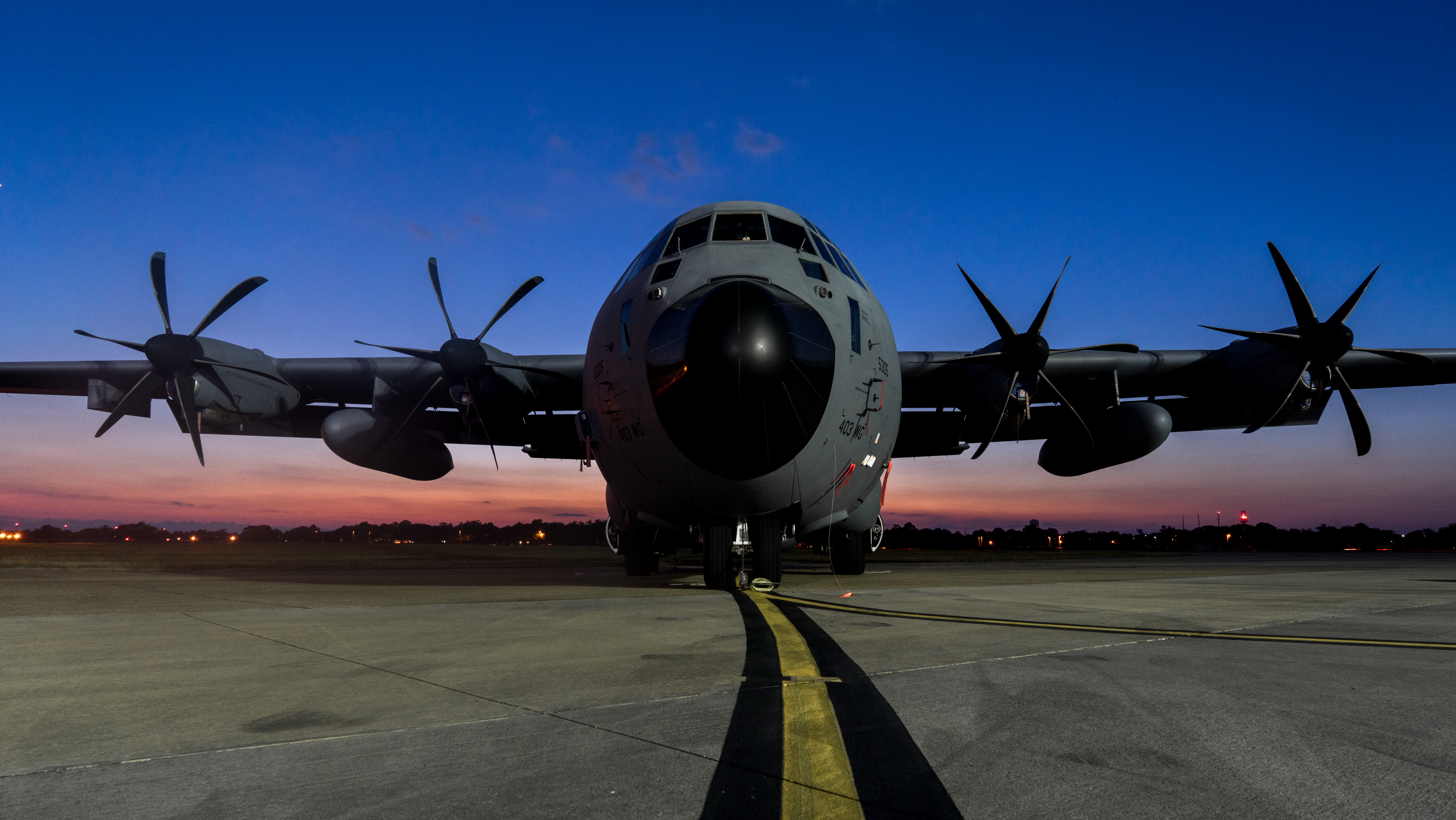 A C-130J is parked on the flightline with a sunset in the background.